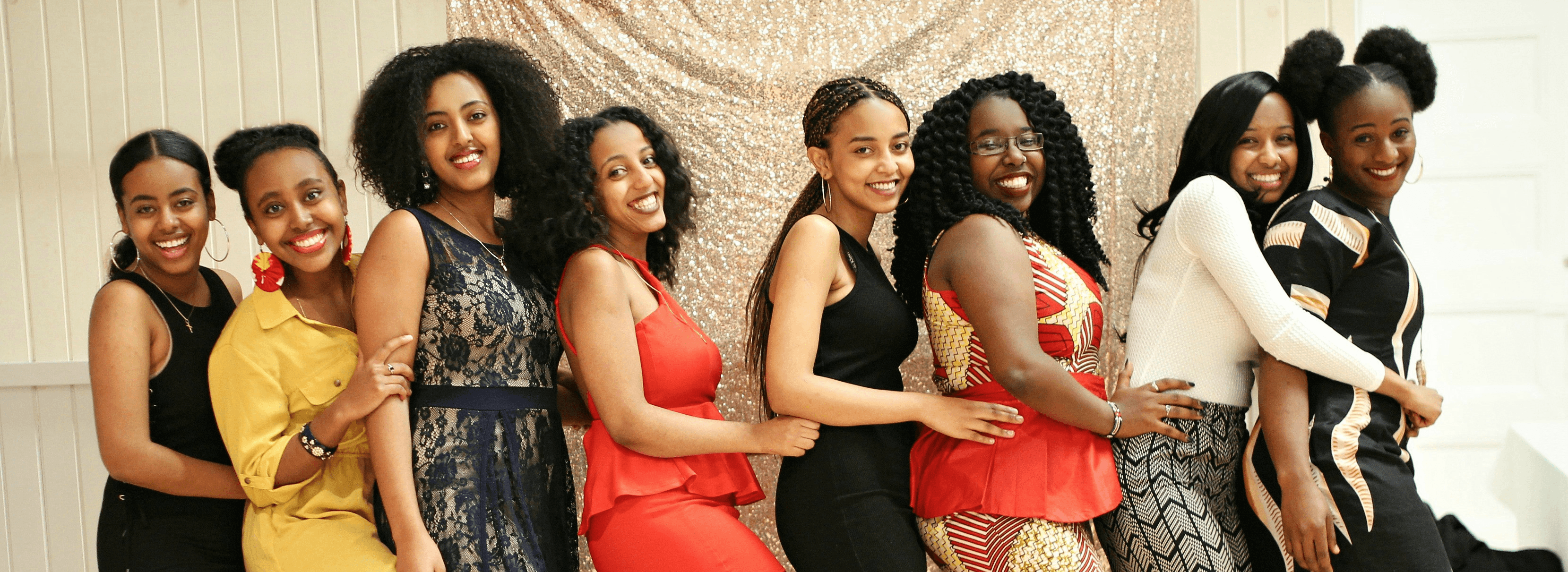 Eight joyful young women pose in a line against a shimmering gold sequined backdrop.