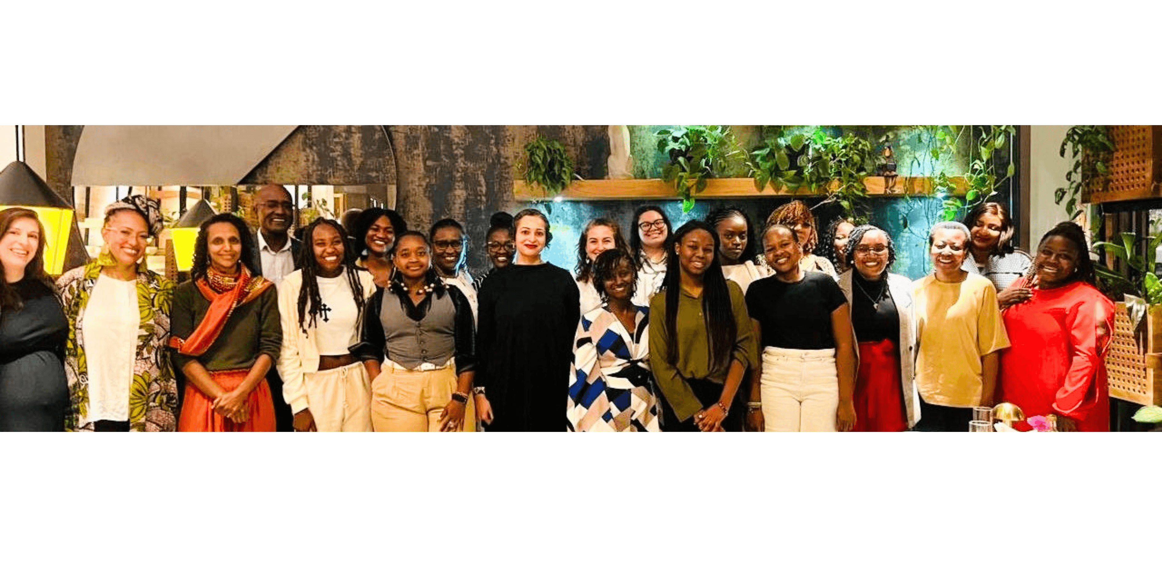 Diverse group of smiling women posing together in an indoor setting with lush greenery.