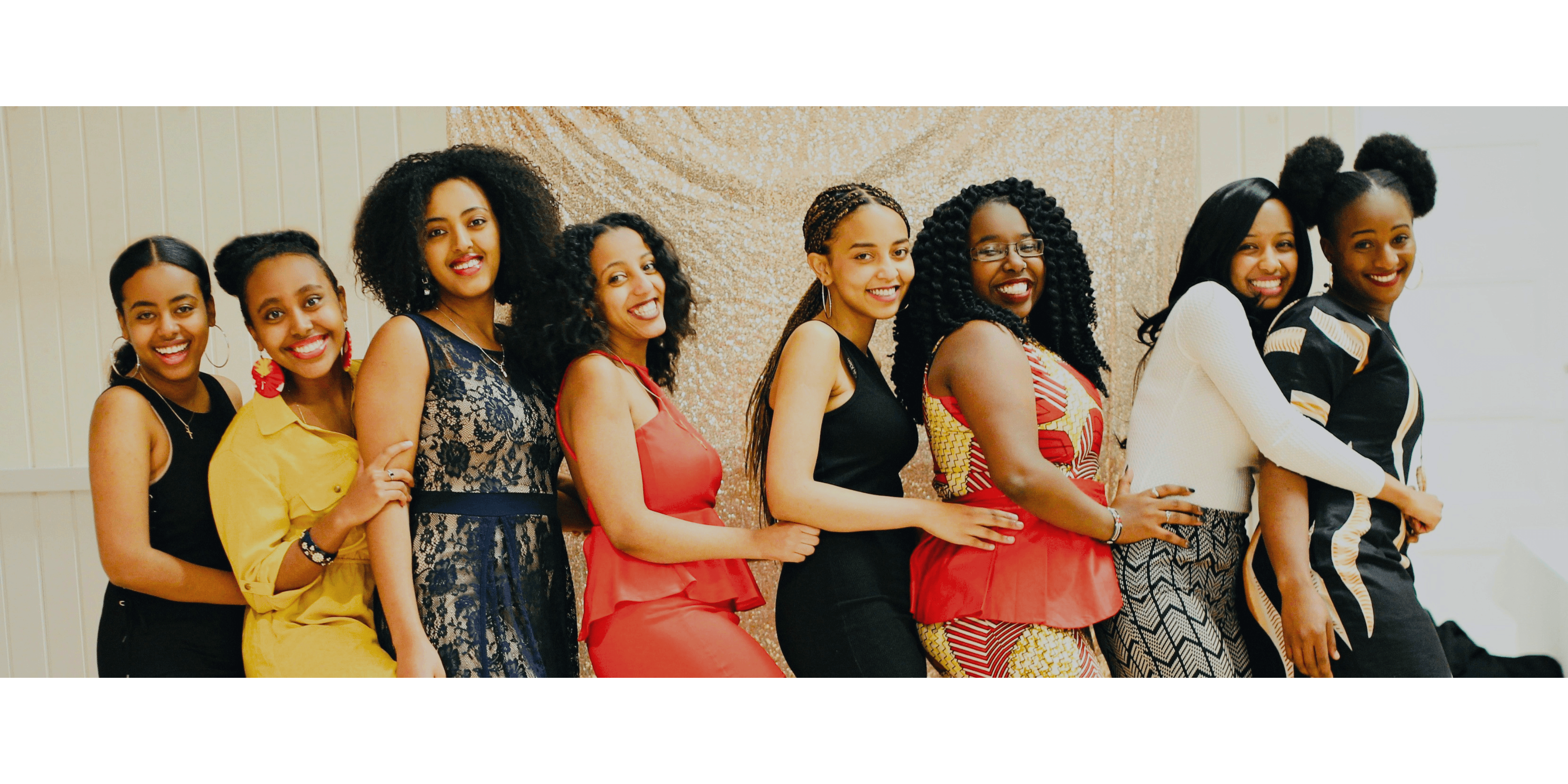 Eight smiling young women in vibrant attire pose in a line against a gold sequin backdrop.