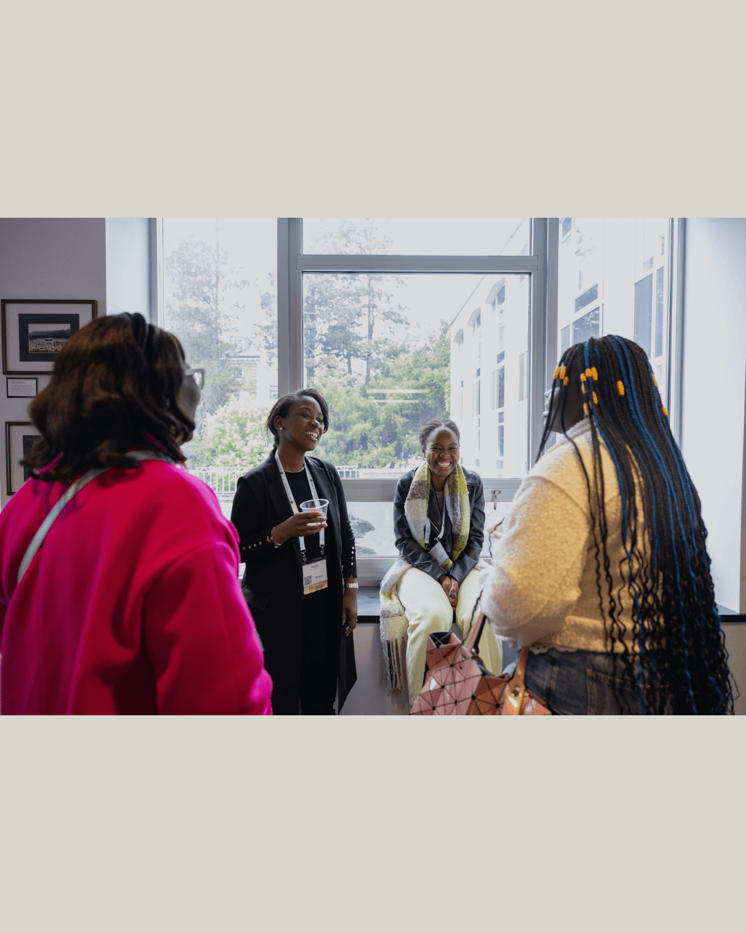 Four Black women smiling and chatting near a window at a professional networking event.