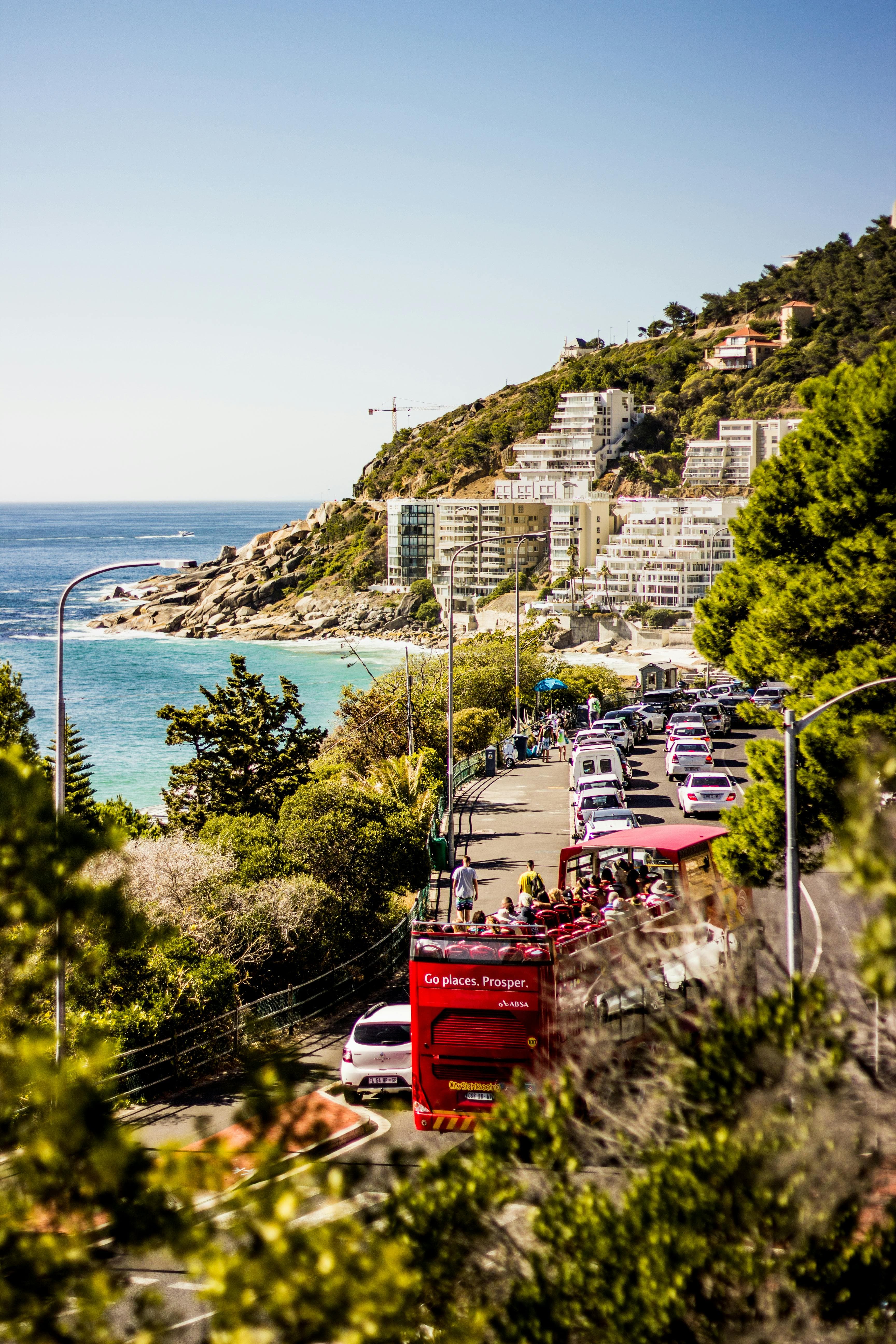 Red open-top tour bus on a coastal road with white buildings and blue ocean.