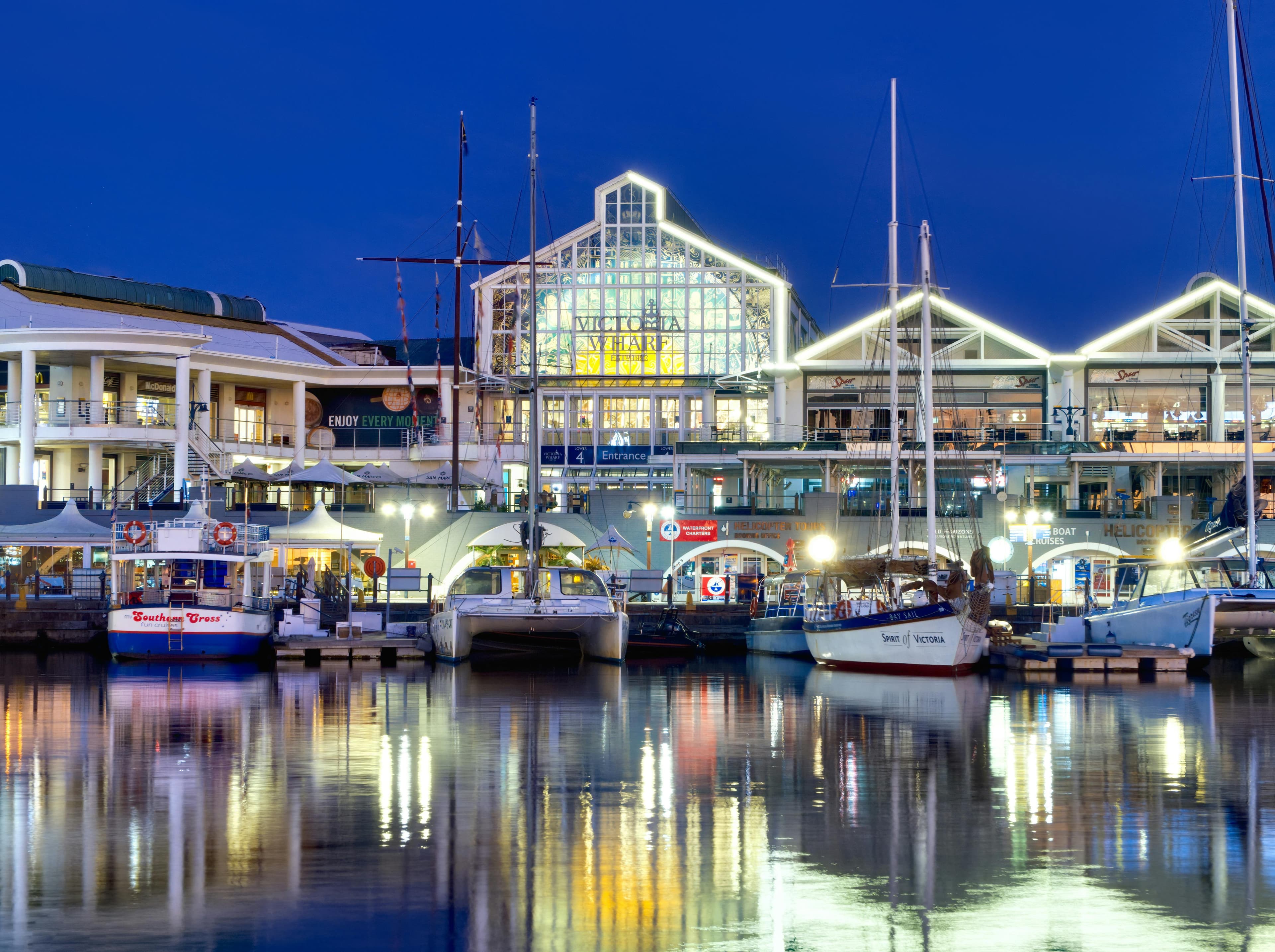 Boats docked at the illuminated Victoria Wharf at night with colorful reflections in the water.