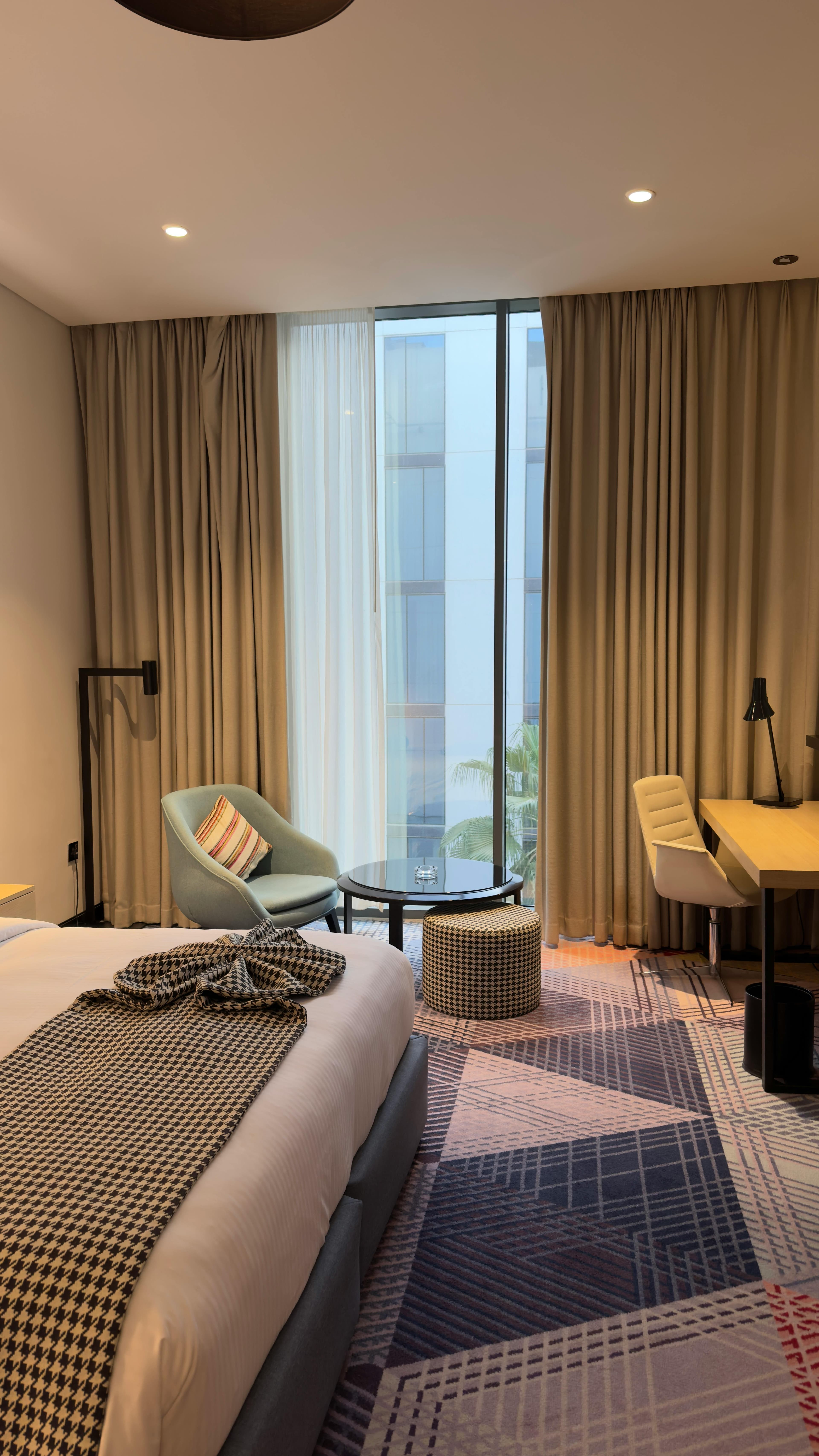 Modern hotel room with a geometric rug, blue armchair, and desk near large windows.
