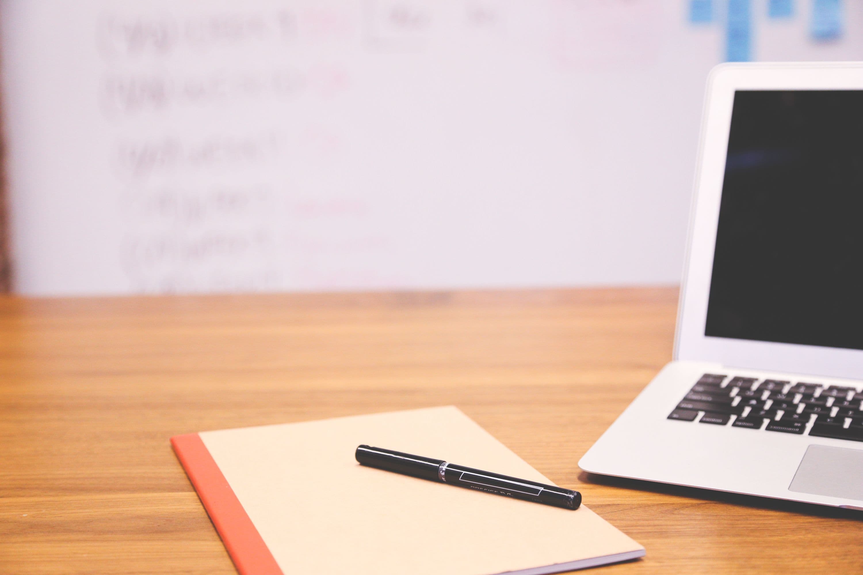 Silver laptop and a notebook with a black pen on a wooden office desk.