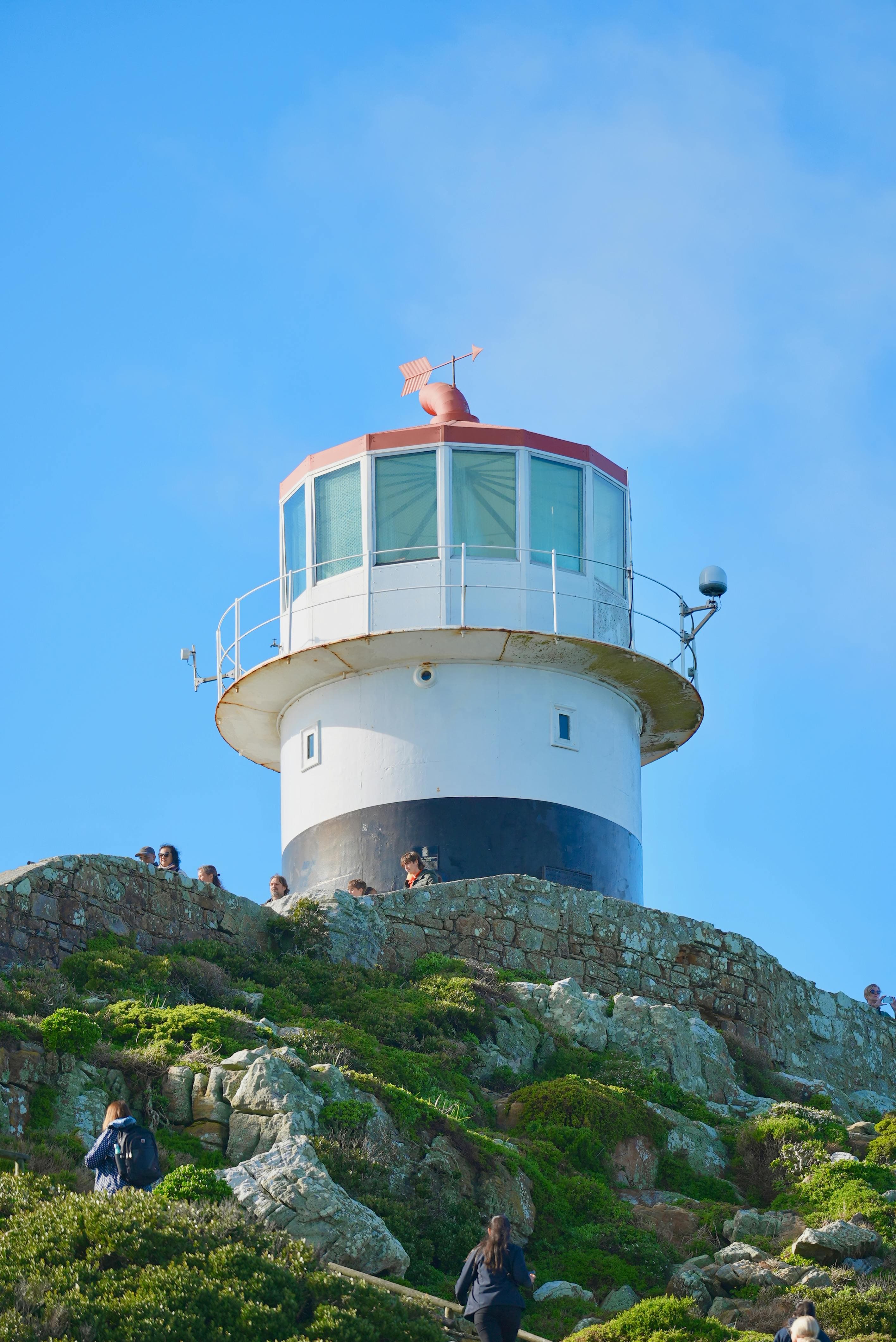 White and black lighthouse with a red top stands on a rocky, green hillside.